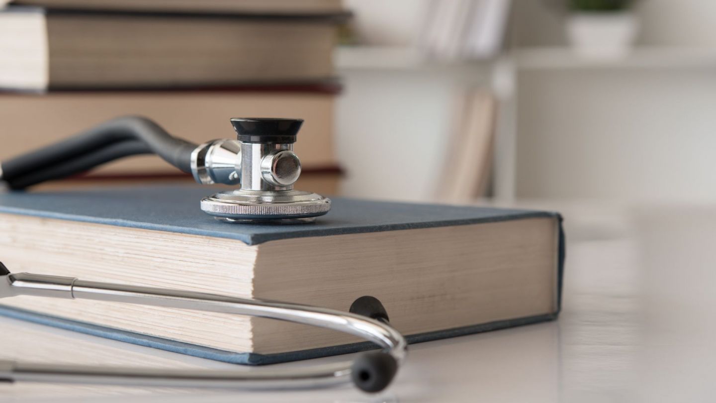 Bir masanın üzerinde bir tıp hukuku ders kitabı ve bir stetoskop var. Arka planda masanın üzerine yığılmış daha fazla kitap var.