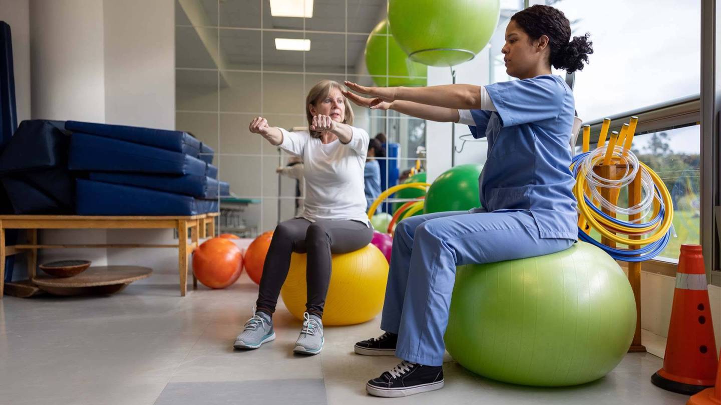 Frau auf einem Gymnastikball macht unter Anleitung einer Physiotherapeutin eine Übung.