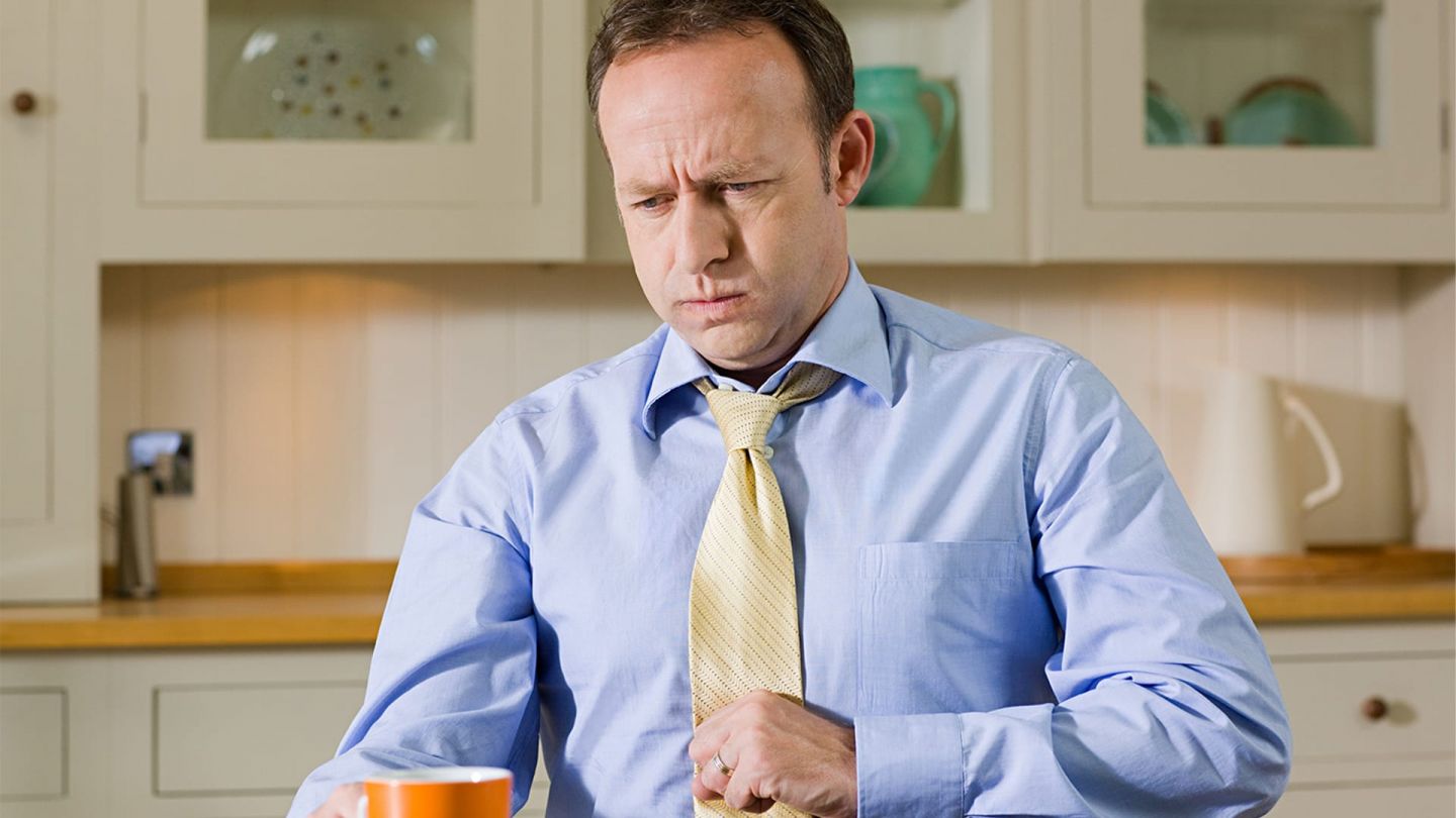 Functional dyspepsia (indigestion): man in suit and tie sitting at the kitchen table. An empty plate and coffee cup in front of him. He is holding his stomach as he looks as if he is about to belch painfully. His face is tense.