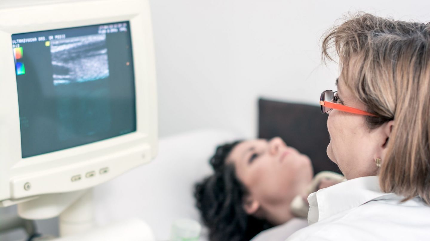 Schilddrüsenkrebs: Eine Patientin liegt auf einer Krankenhausliege. Vor ihr sitzt eine Ärztin und führt eine Ultraschalluntersuchung ihrer Schilddrüse durch. Die Ärztin beobachtet das Ultraschallbild auf einem Monitor.