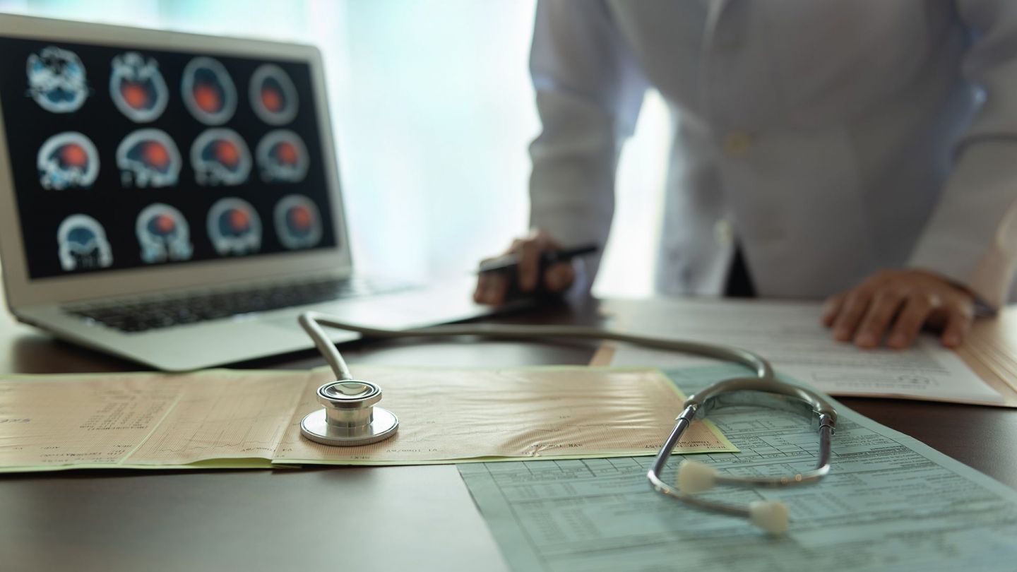 Ein Arzt analysiert Berichte, die neben einem Stethoskop auf einem Tisch liegen. Ein Bildschirm zeigt Bilder eines gescannten Schädels und Gehirns.