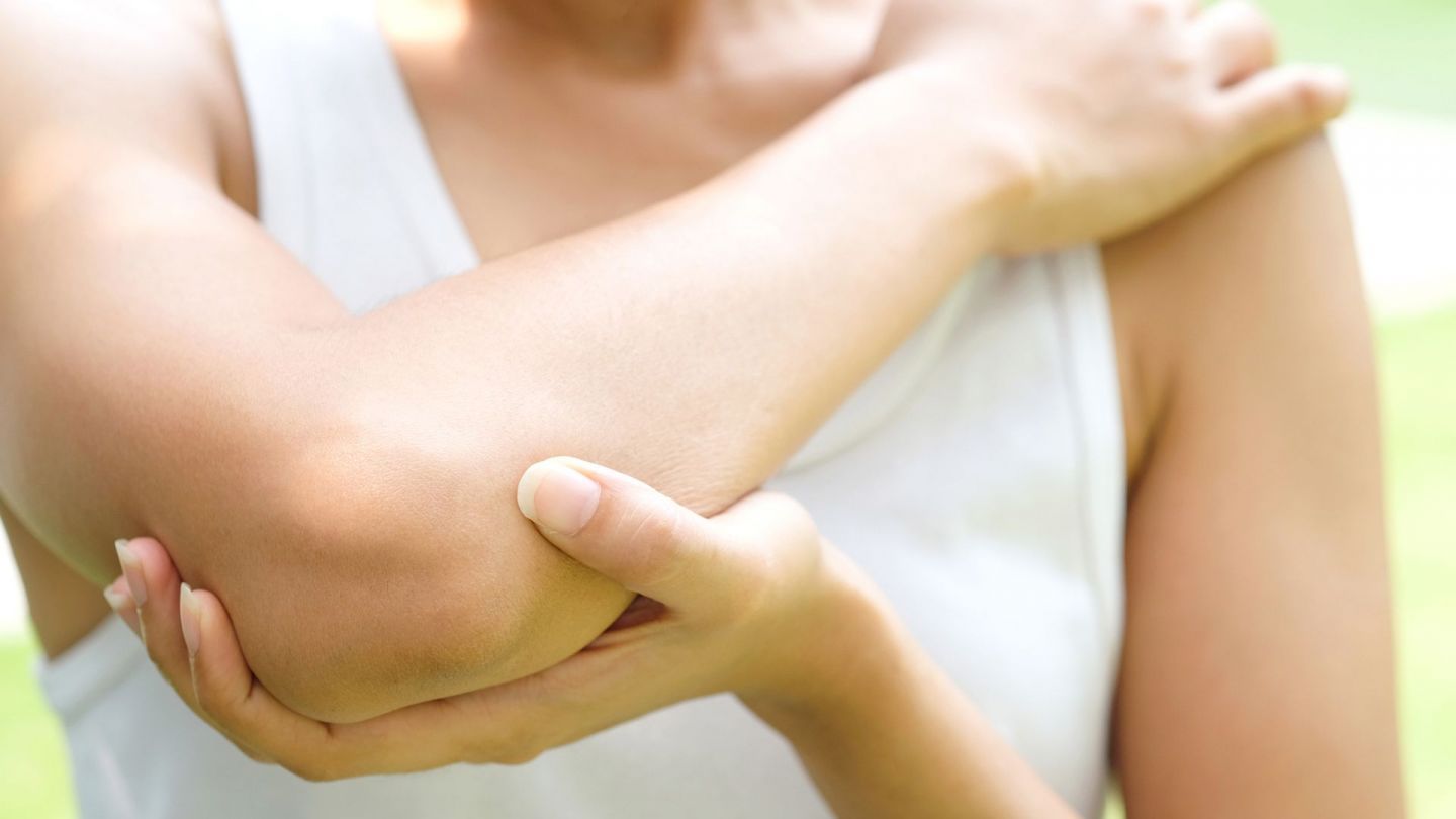 Schleimbeutelentzündung: Eine junge Frau mit sportlicher Kleidung streckt einen Arm nach oben und winkelt den Ellenbogen des Arms an. Mit der anderen Hand stützt sie den Ellenbogen.
