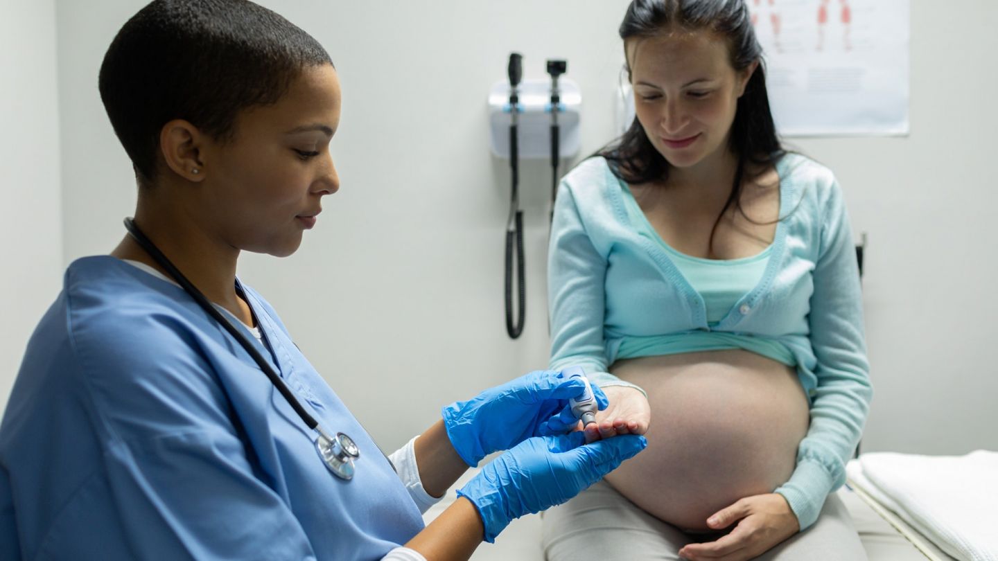 Gestational diabetes: pregnant woman sitting on a treatment table. A woman is standing next to her wearing a blue lab coat and surgical gloves. She is holding out a measuring device to the pregnant woman’s outstretched hand.