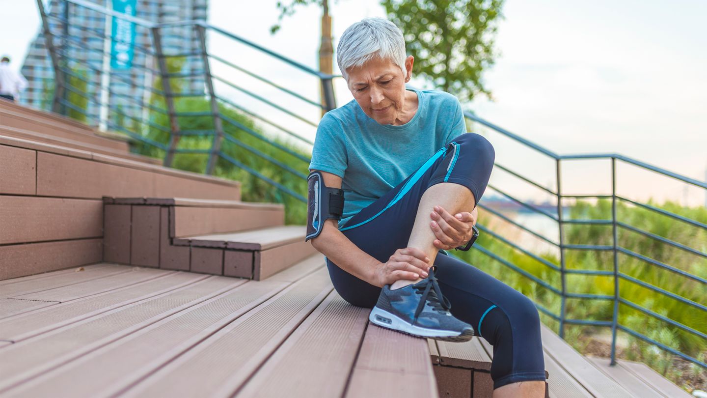 Tendinitis: older woman in sports clothing sitting on steps in the open air. She is holding her ankle and grimacing with pain.