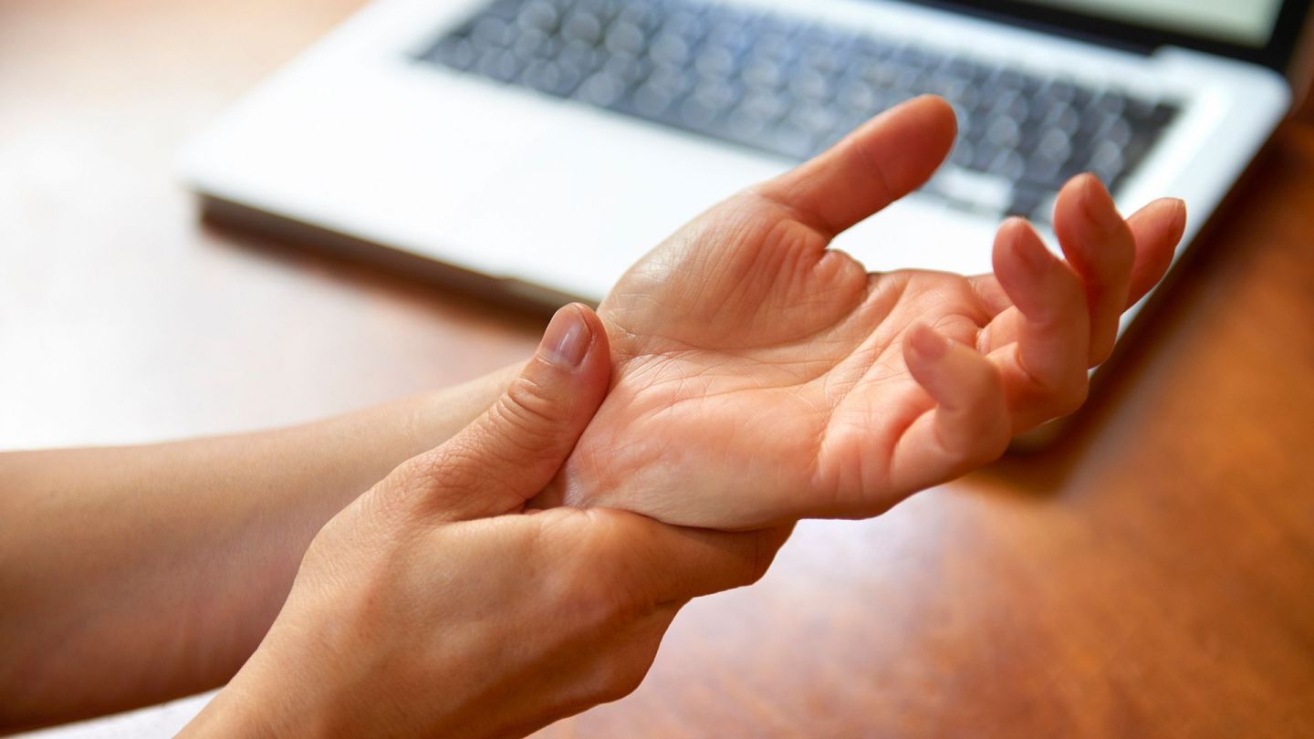 Tenosynovitis: woman sitting at a desk with a laptop, she is clearly working. The woman is clutching her left wrist with her right hand. She is pressing her thumb down on her wrist joint.