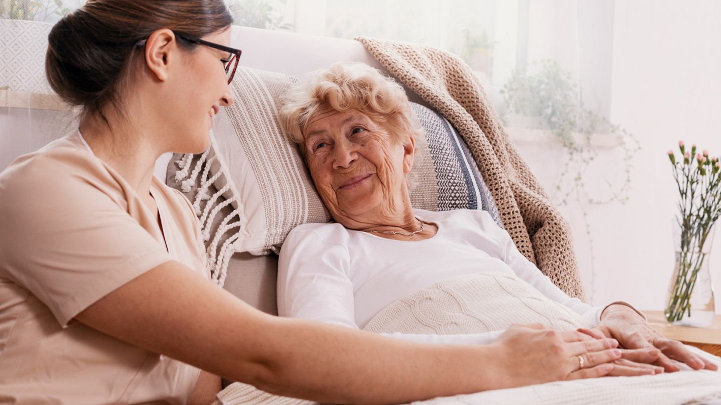 Редкие виды рака: пожилая женщина лежит в постели. Медсестра сидит рядом с ней и держит ее за руку. Обе улыбаются друг другу.