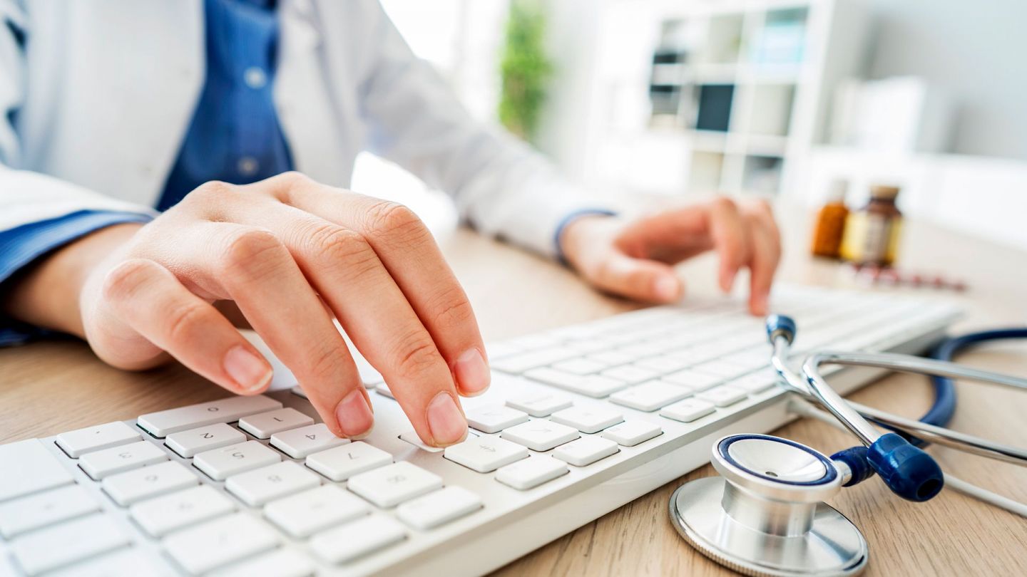 Second opinion: doctor sitting at a desk using the fingers of both hands to type on a computer keyboard. There is a stethoscope next to the keyboard.