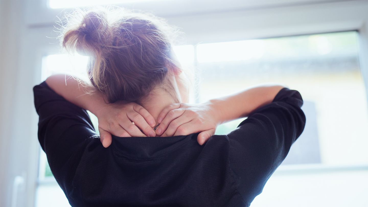 Stress: woman with hair tied up standing in front of the window stretching her arms up and pressing down on her tense neck with her hands. She is twisting her head towards her shoulder to stretch her neck.