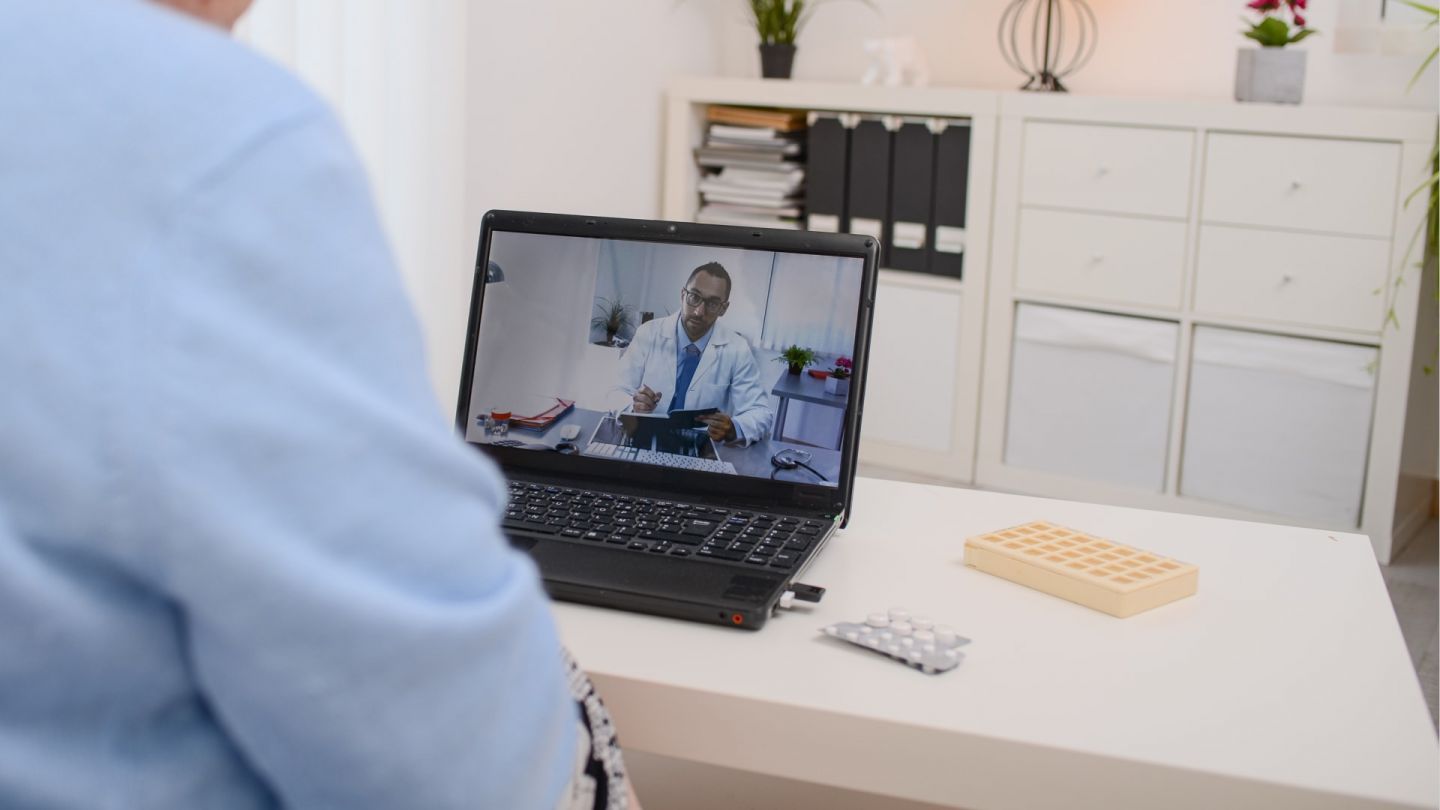 Telemedicine: woman sitting in a living room with a table on which a laptop is standing. She sees a doctor looking at her on the screen. The doctor is clearly looking into a screen ready to talk to the woman.
