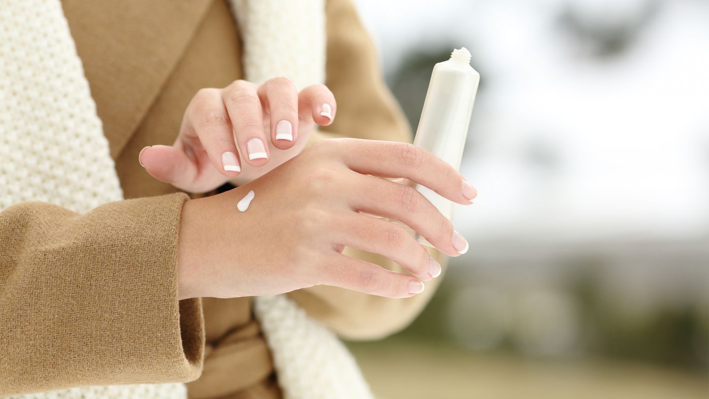 Dry skin: woman wearing a winter coat. She is holding white cream in her hand. She is applying moisturizing cream with one hand to her other hand.