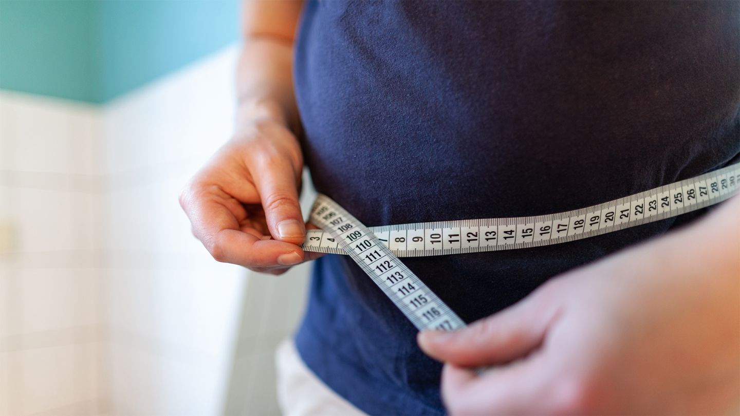 Übergewicht: Ein Mann misst seinen Bauchumfang mit einem Maßband, das er mit beiden Händen vor dem Bauch zusammenhält.