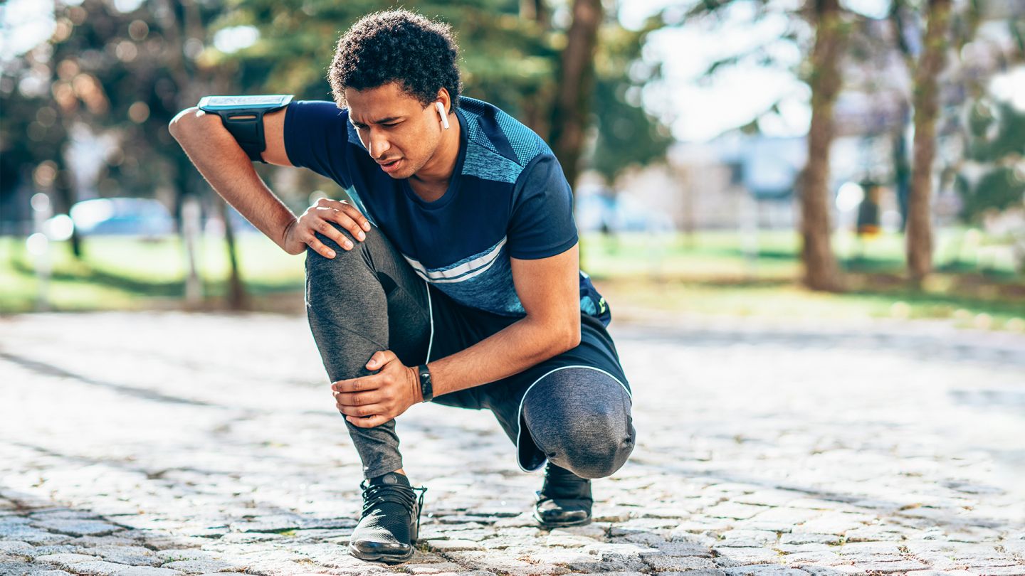 Anterior knee pain: young man in sports clothing kneeling down in the open air. He is holding his knee and grimacing. He appears to have stopped in the middle of a training run because of pain.