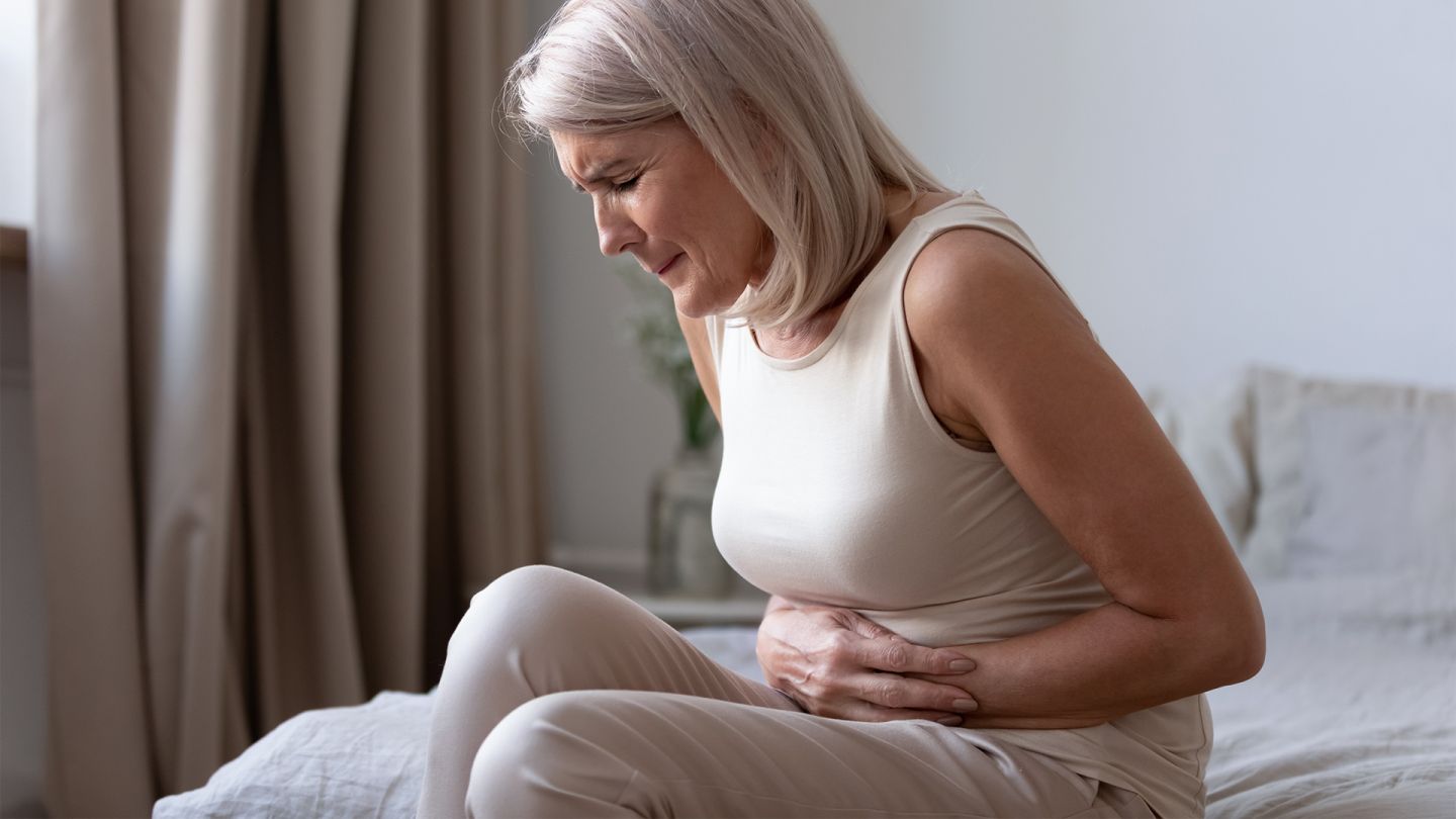 Duodenal ulcer: older woman sitting on the bed. She is holding her belly and drawing up her legs, grimacing with pain.
