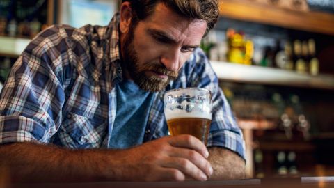 Alkohol: Ein Mann sitzt alleine in einer Bar, lehnt sich mit beiden Armen auf den hölzernen Tresen, hält ein Bierglas in der Hand und schaut in den Glas.