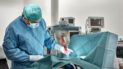 Острый миелоидный лейкоз у взрослых: хирург, одетый в хирургический костюм, стоит в операционной. Перед ним на операционном столе лежит накрытый простыней пациент. С помощью шприца у пациента берется образец костного мозга. Врач контролирует процесс.