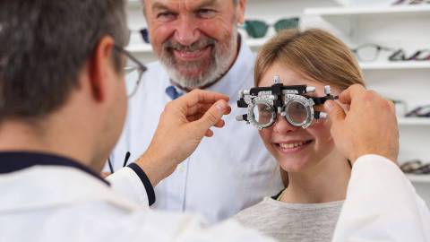 Bir gözlükçü veya göz doktoru, göz testi için bir kıza ölçüm gözlüğü takıyor. Yaşlı bir adam gülümseyerek yanında duruyor.