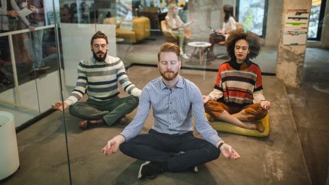 Workplace health promotion: 3 people sitting on the floor of a room with glass walls in an office building. They are sitting cross-legged with their arms resting on their knees. They look as though they are meditating during working hours.