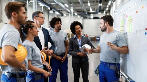 Workplace health management: 5 people in a storage depot receiving instructions. 4 people are wearing protective clothing, 2 are dressed in formal office clothes. They are all listening closely.