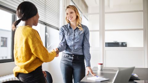 Workplace Integration Management: two women standing at an office desk shaking hands in agreement after a successful business meeting.