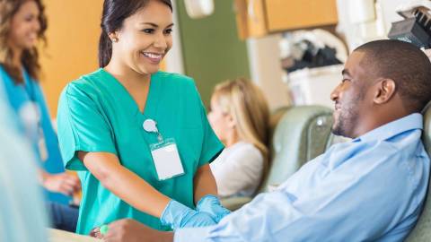 Gülümseyen bir sağlık görevlisi tedavi sandalyesinde oturan bir kan bağışçısıyla ilgileniyor.