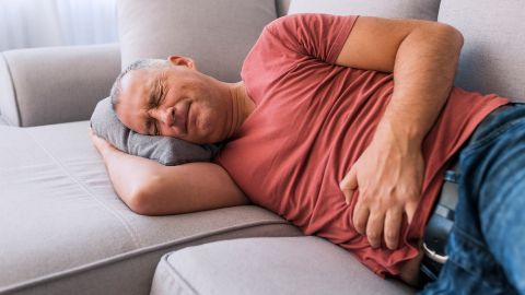 Botulism: man lying on a sofa holding his stomach with his left hand. He has his eyes closed and he is grimacing in pain.