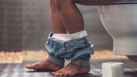 Campylobacter infection: man sitting on the toilet with his trousers down. Only his legs are visible. There is a roll of toilet paper by his feet.