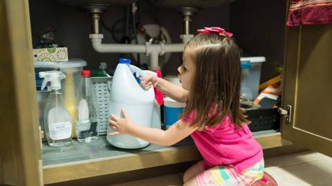 A young girl kneels in front of a shelf of cleaning agents and takes out a large bottle.