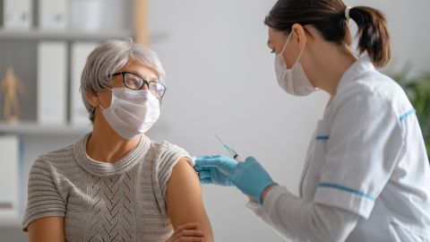COVID-19 aşısı: Bir doktor yaşlı bir bayanın koluna aşı enjekte ediyor. Her ikisi de ağız ve burun koruması kullanıyor.