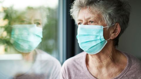COVID-19: Eine ältere Frau schaut aus dem Fenster. Über Mund und Nase trägt sie einen Mundschutz.
