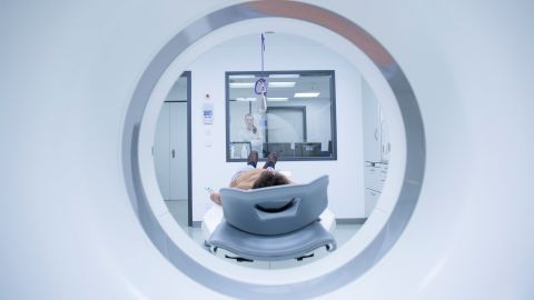CUP-Syndrom (Krebs bei unbekanntem Primärtumor): Ein Patient liegt vor einer MRT-Röhre. Im Hintergrund steht eine Ärztin hinter einer Glasscheibe und beobachtet den Patienten.