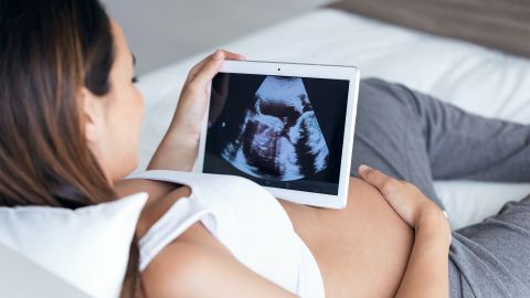 E-anne kartı: Gebe bir kadın, doğmamış çocuğunun ultrason görüntüsüne bir tablette bakıyor. Tablet çıplak karnının üzerinde duruyor, rahat ve memnun görünüyor.