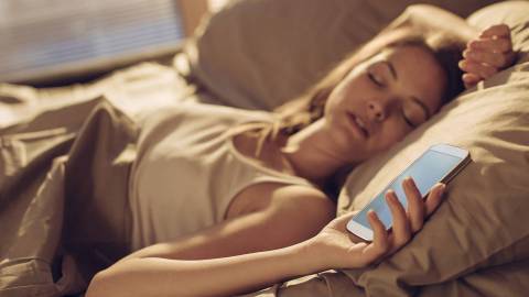 Bir kadın yatakta uyuyakalmış ve elinde bir akıllı telefon tutuyor.