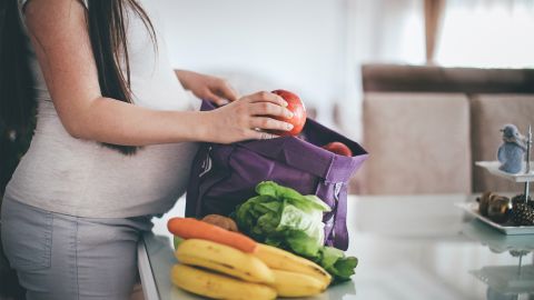 Pregnant woman standing in the kitchen unpacking groceries. She has bought lots of loose, unpackaged fresh fruit and vegetables.