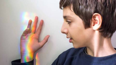A boy is holding his hand up to a white wall. The rainbow colors of refracted sunlight are reflected on his hand.