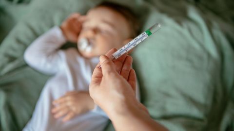 High temperature (fever) in children: one hand holding a thermometer, baby lying in a bed in the background.