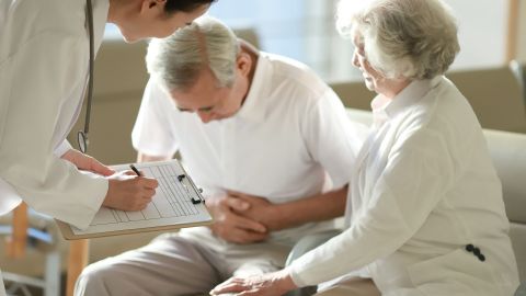 Gallstones: older man sitting on a couch with his chest bent forward pressing both hands to his abdomen. A woman is sitting next to him touching his left leg with one hand. A doctor is standing in front of them taking notes.