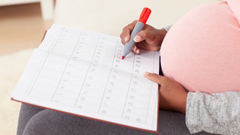 Geburtsvorbereitung: Eine hochschwangere Frau markiert sich mit einem roten Marker den geplanten Geburtstermin in einem großen Kalender.