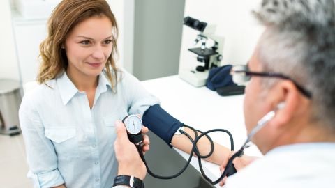 Gesundheits-Check-Up: Eine Frau sitzt einem Arzt gegenüber. Sie trägt eine Manschette um den Arm, ihr Blutdruck wird gemessen.