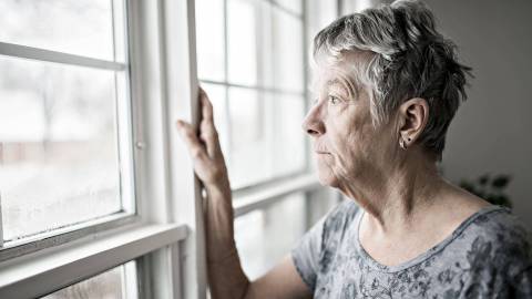 Eine ältere Frau lehnt am Fenster und schaut betrübt nach draußen.