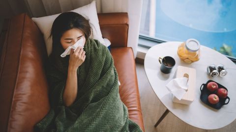 Grippe: Eine junge Frau sitzt in einem Sessel. Ihr Oberkörper ist bis unters Kinn mit einer Decke eingehüllt. Mit einer Hand hält die Frau ein Taschentuch an ihre Nase.