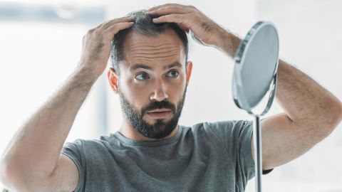 Hair loss: young man gazing at himself in the mirror. He has three-day-old stubble and is examining areas where the hair on his head is thinning.