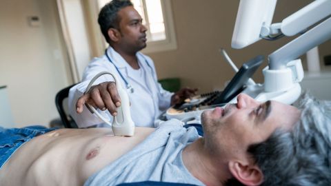 Heart valve disease: middle-age man lying on his back on a treatment table. A doctor is standing next to him. He is holding a scanner to the man’s ribcage in one hand. The doctor is giving him an ultrasound examination.