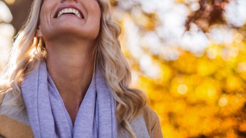 Glücksgefühle durch Hormone: Eine Frau, die ein Handtuch um den Hals trägt, legt ihren Kopf in den Nacken und lächelt. Im Hintergrund Laub, Äste und Bäume. 