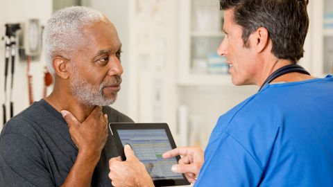 Kehlkopfkrebs: Ein älterer Mann fasst sich mit der linken Hand an den Hals. Ihm gegenüber steht ein Arzt, der ein Tablet in beiden Händen hält.