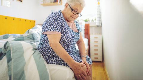 Пожилая женщина сидит на кровати и держится за больное колено.