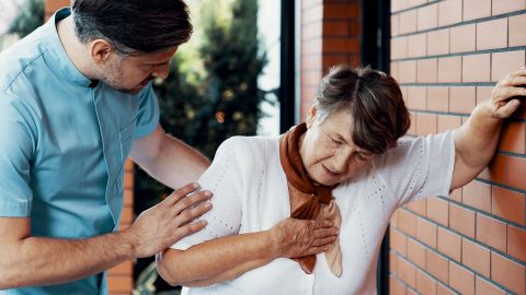 Coronary heart disease: older woman leaning against the wall of a house, clutching her chest with her other hand. The woman appears to be exhausted. A younger man is standing next to her and supporting her shoulder.