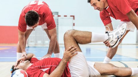 Çapraz bağ yırtılması: Bir futbolcu yerde yatıyor, yüzü acıyla çarpılıyor, bir eliyle başını, diğer eliyle bacağını tutuyor. Bir takım arkadaşı yanında duruyor ve yaralı kişinin ayağını destekliyor.