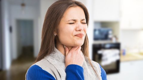 Воспаление миндалин: женщина держится рукой за шею, у нее искажено лицо и зажмурены глаза.