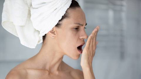 A woman with a towel wrapped around her head coughs into her open hand to check her breath.