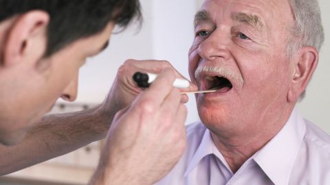 Oral cancer: doctor examining an older man’s mouth. The man has his mouth open wide. The doctor is pressing his tongue down with a wooden tongue depressor and shining a light into his mouth.